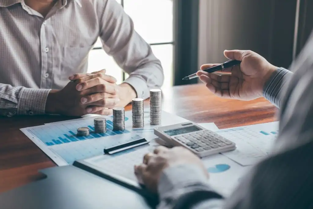 Two people at a wooden desk with stacked coins, a calculator, and a printed bar chart