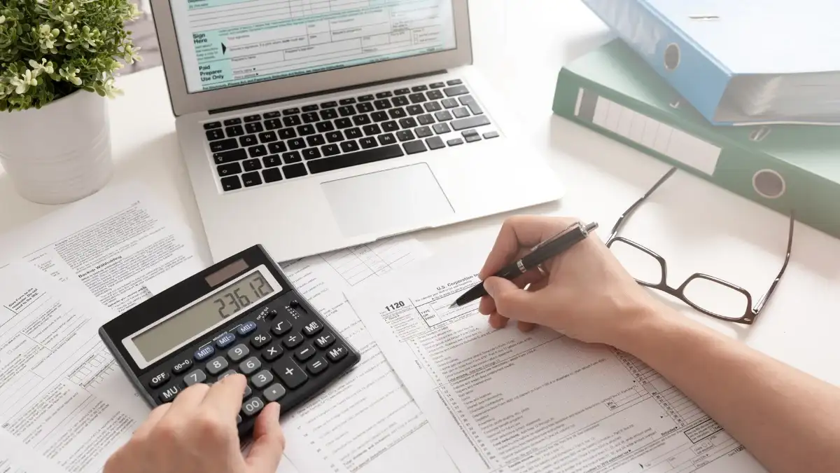 Hands with a calculator and a printed form beside a laptop and binders